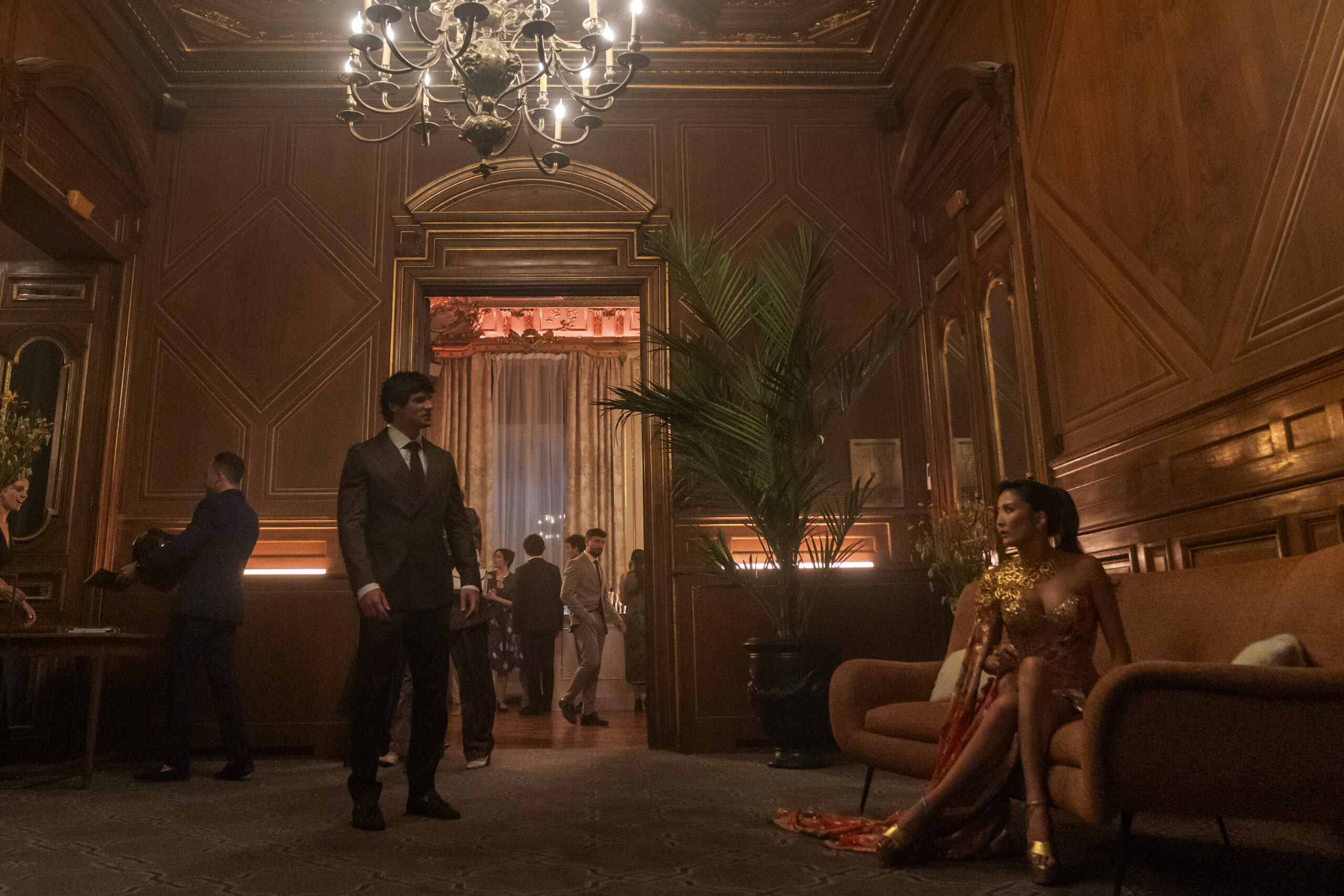 "A man in a suit stands in an elegant, wood-paneled lounge with a chandelier, while a woman in a gold dress sits on a couch; other people are visible in the background socializing at a formal event."