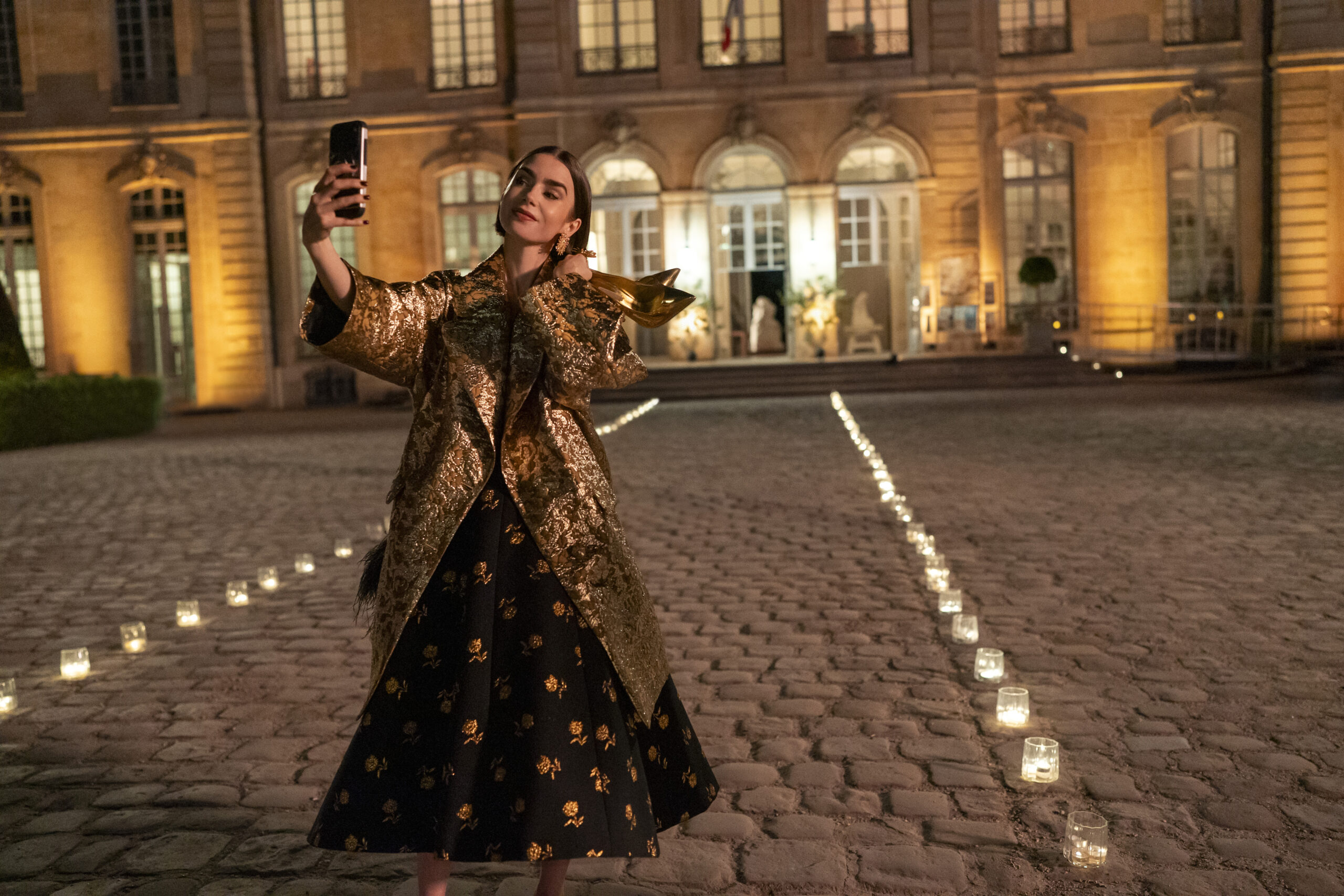 "Woman in a gold coat and black dress takes a selfie with her phone at night in a candlelit courtyard outside an elegant, well-lit building with large windows and cobblestone ground."