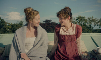 "Two women in historical clothing sit outside on a rooftop, smiling at each other with greenery and blue sky in the background. Pillows and a cup are nearby, creating a relaxed, intimate setting."