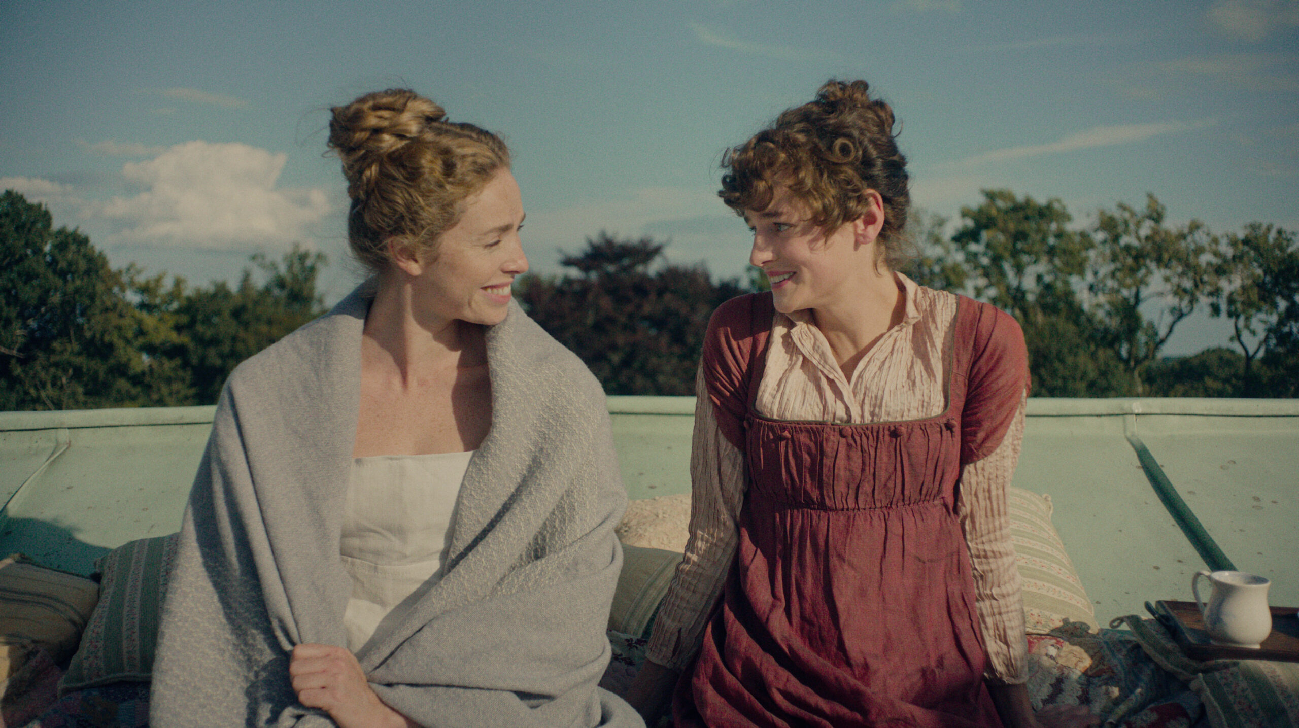 "Two women in historical clothing sit outside on a rooftop, smiling at each other with greenery and blue sky in the background. Pillows and a cup are nearby, creating a relaxed, intimate setting."