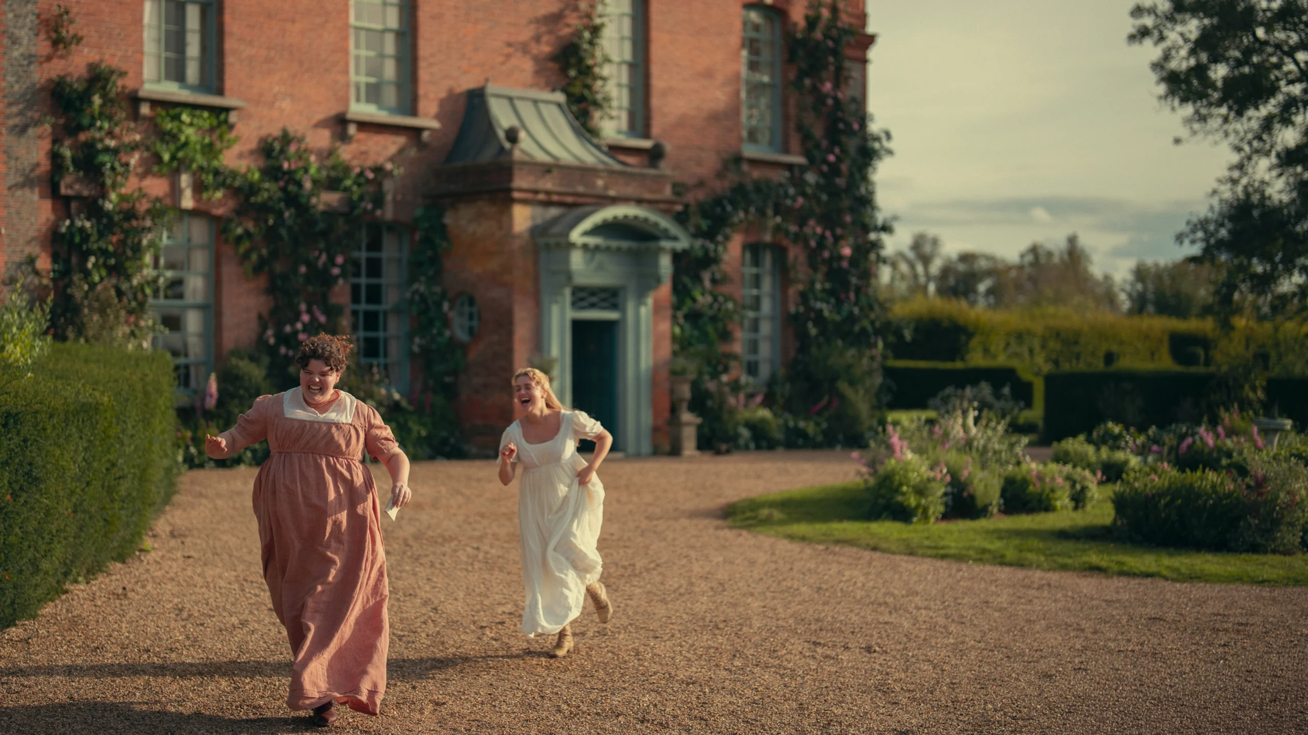 "Two women in period dresses run joyfully on a gravel path in front of a large brick house with ivy and flowers, surrounded by hedges and gardens under a partly cloudy sky."