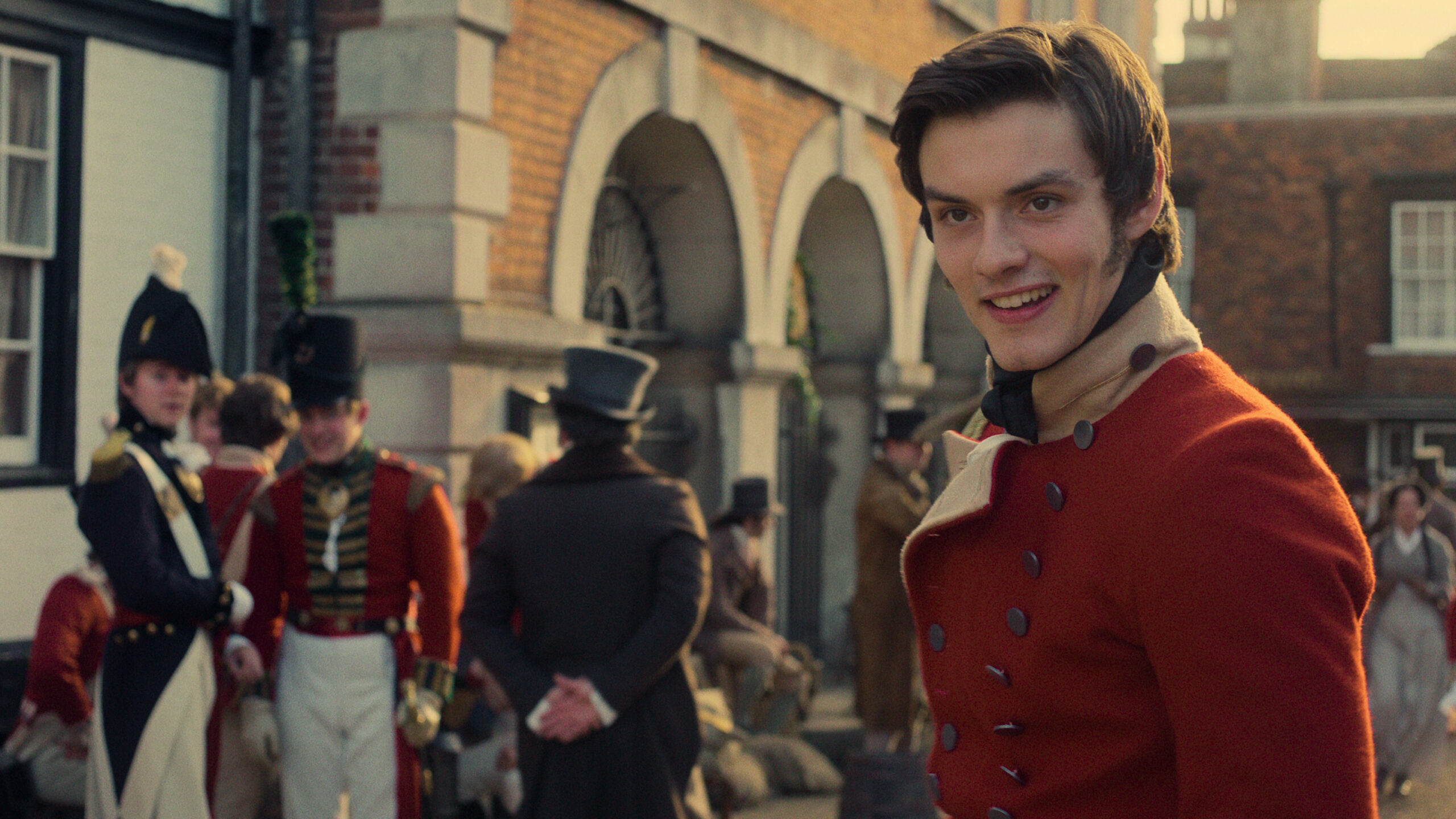 "Young man in red historical military uniform smiles in foreground, with other soldiers and townspeople gathered outside a brick building with arches in a period village setting, evoking a historical or Regency-era atmosphere."