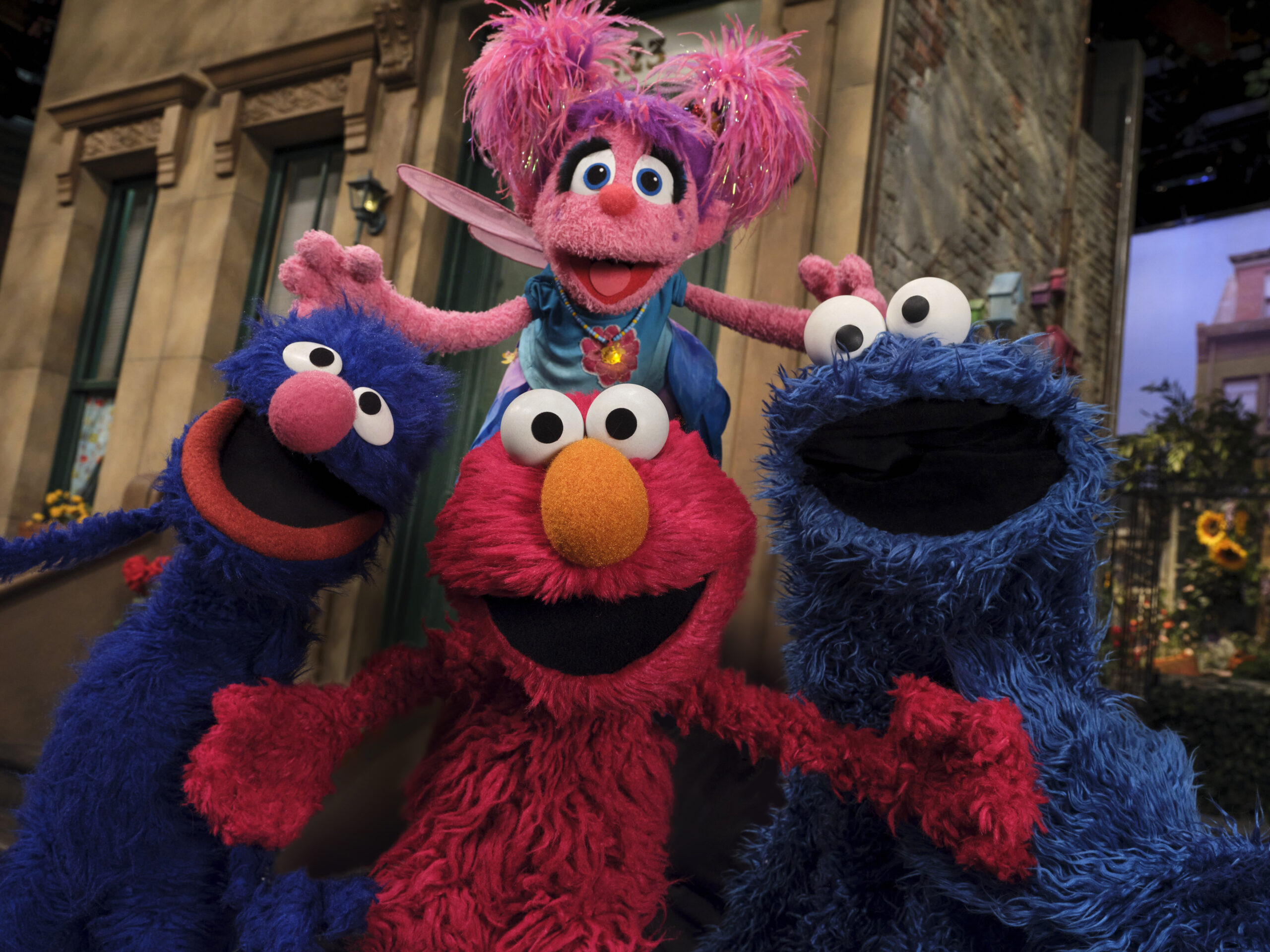 "Four colorful Sesame Street puppet characters are smiling and posing together outside in front of classic city brownstone apartment buildings on a sunny day."